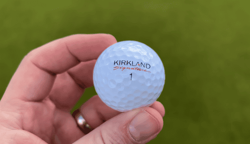 hand with ring holds golf ball with kirkland signature on it in front of the grass