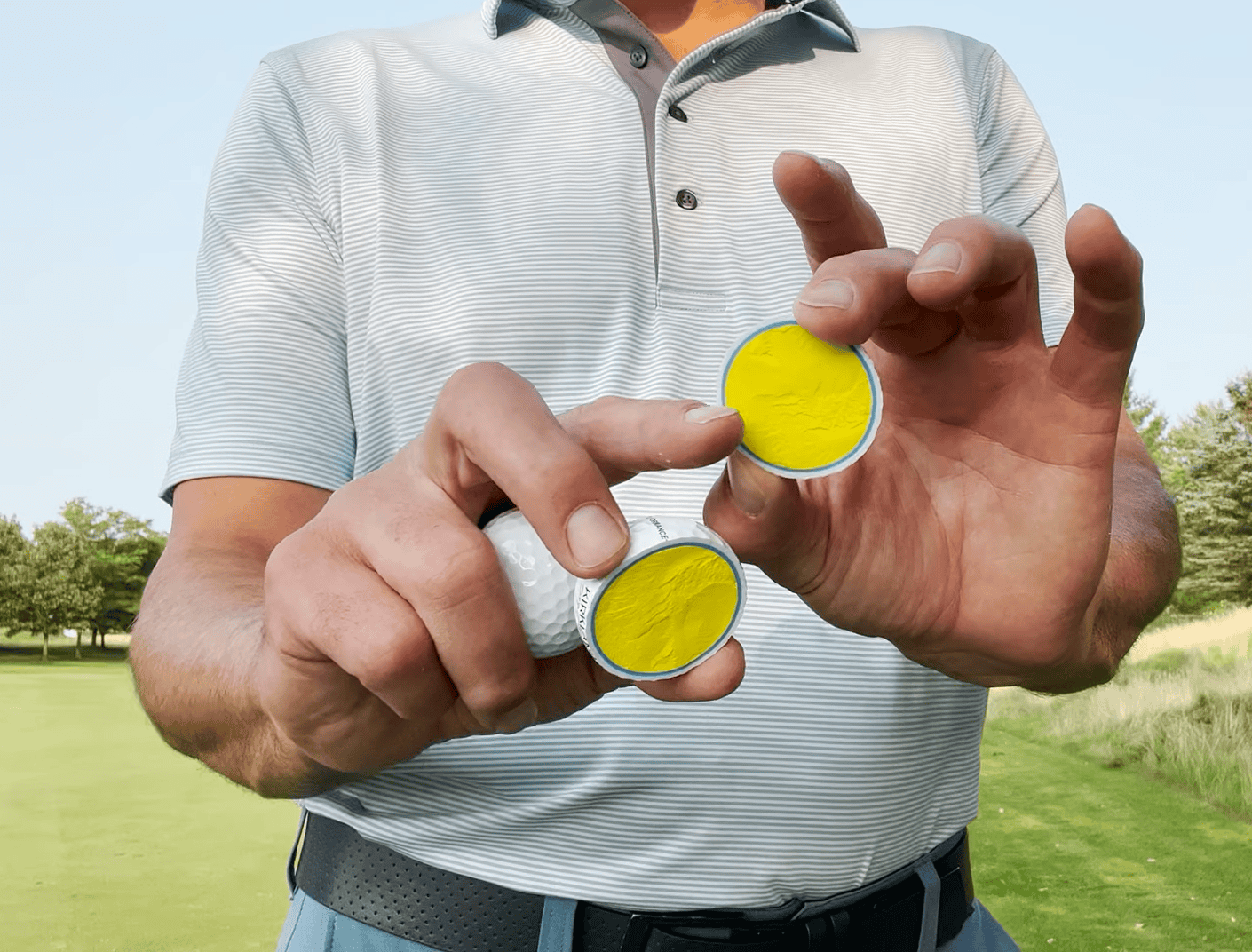 man in polo shirt and jeans showing cut golf ball, the field with trees behind