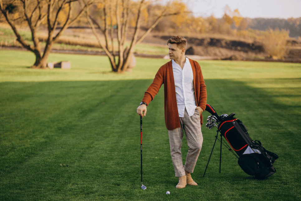 man in white shirt, brown jacket and beige pants holds golf club and bag with golf equipment on field